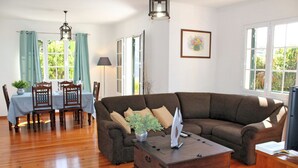 A living room with a brown sectional sofa, a wooden coffee table, and a dining area with a table and chairs. - Perched above village, ocean views | Jardim Mar (Ponta do Sol)
