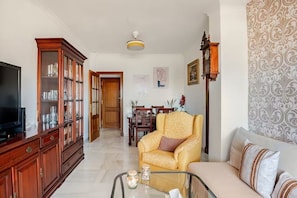 Una sala de estar con televisor, una mesa de centro de vidrio, un sofá, un papel tapiz estampado y un mueble de madera.