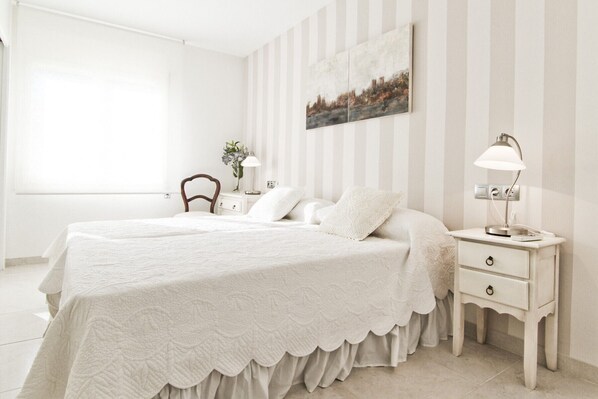 A bedroom with two beds, a nightstand with a lamp, a chair, and a wall-mounted painting.