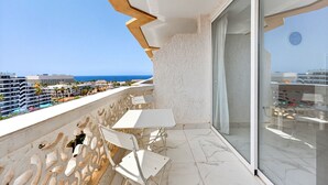 Uma varanda com mesa e cadeiras brancas, porta de vidro e vista para o oceano e edifícios.