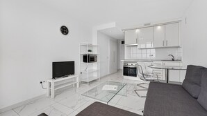 Uma sala de estar moderna com televisão de tela plana, mesa de centro de vidro, sofá cinza e área de cozinha com armários e eletrodomésticos brancos.