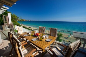 A wooden table set with plates, cups, and a fruit bowl, overlooking a beach with clear blue water and a distant shoreline. - Infinity Beach House (Kefalonia)