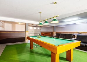 Une table de billard recouverte d’un feutre vert, une table en bois avec un bord jaune, et deux lampes suspendues vertes.