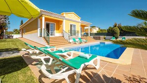 Una casa amarilla con piscina, sillas de descanso verdes y un porche cubierto.