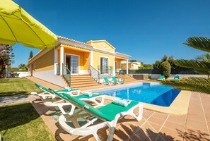 A yellow house with a swimming pool, green lounge chairs, and a covered patio area.