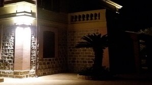 Un bâtiment doté d’une façade en pierre, d’une entrée imposante surmontée d’une arche illuminée, et d’un palmier au premier plan.