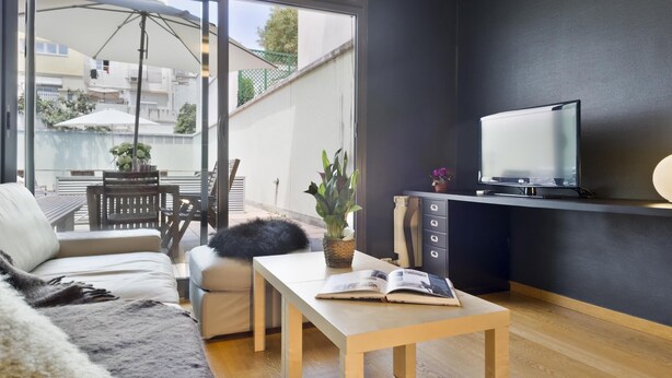 Una sala de estar moderna con un sofá, una mesa de centro, un televisor sobre un mueble negro y un balcón con mesa y sillas.