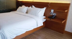 A hotel room with a wooden headboard, a bed with white linens, a bedside table with a phone and power outlets, and a wooden floor.