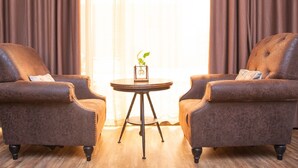 Zwei braune Sessel stehen sich gegenüber, dazwischen ein kleiner runder Tisch. Auf dem Tisch steht eine Pflanze, im Hintergrund ein Fenster mit Vorhängen.