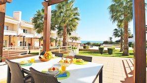 An outdoor dining area with a table set for breakfast, including fruits, pastries, and juice, overlooking a pool and palm trees.