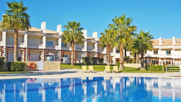 A swimming pool with clear blue water, surrounded by palm trees and a row of white buildings with balconies.