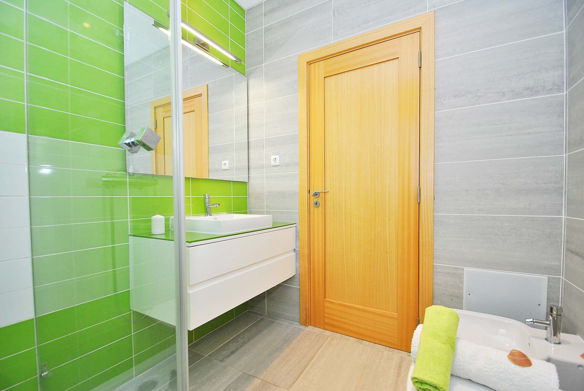 A modern bathroom with a glass shower enclosure, a wooden door, and a white sink with a green countertop.