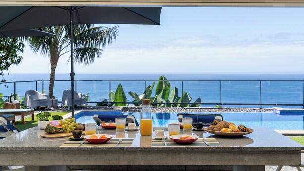 A breakfast setup with a view of the ocean, including a table with plates of food, a bottle of orange juice, and a tropical setting with palm trees and a pool.