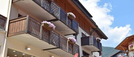 A multi-story building with balconies adorned with flower boxes, a visible storefront with signage, and a stone staircase leading to the entrance.