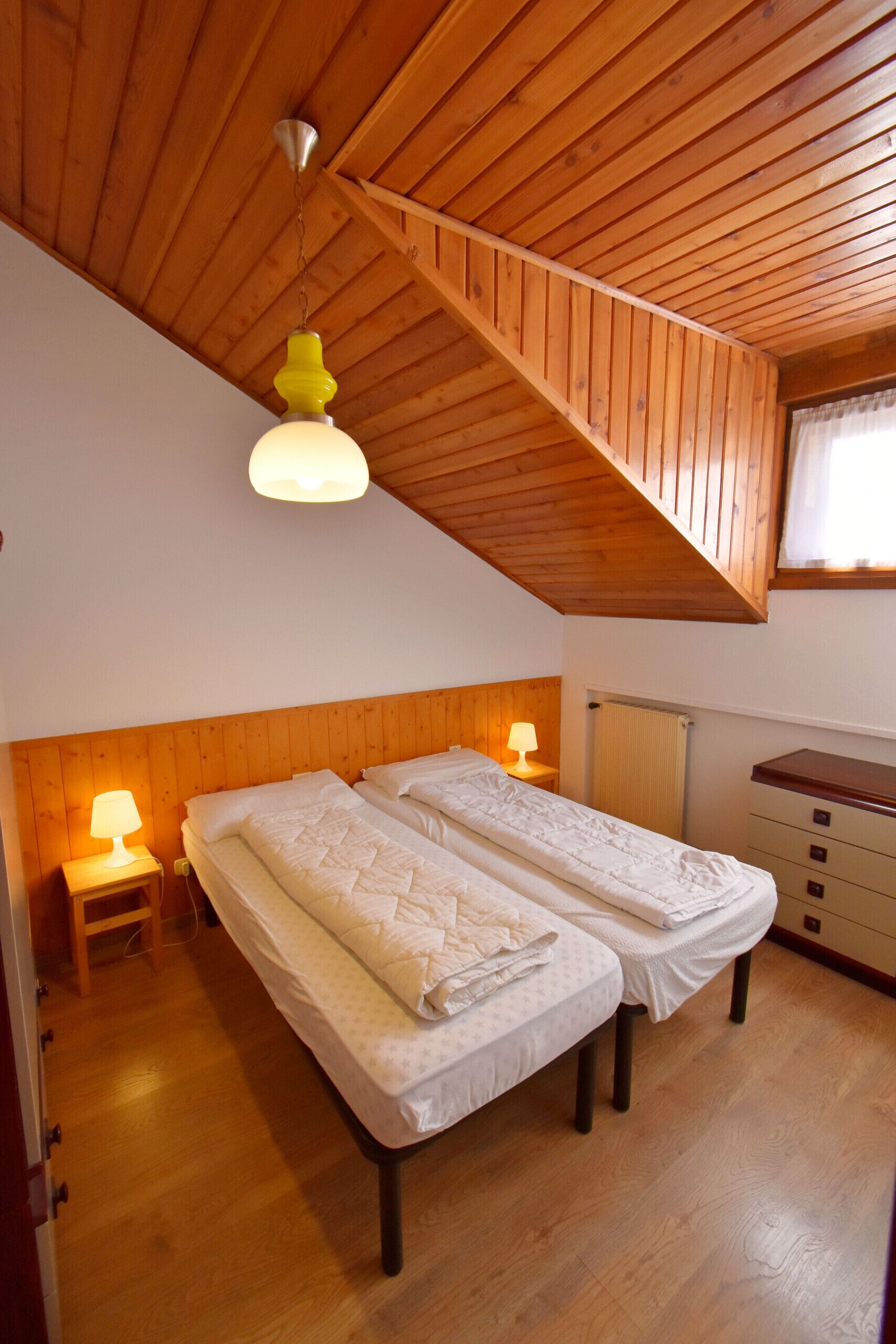 A room with two beds, wooden ceiling beams, a small desk, and a window with curtains.