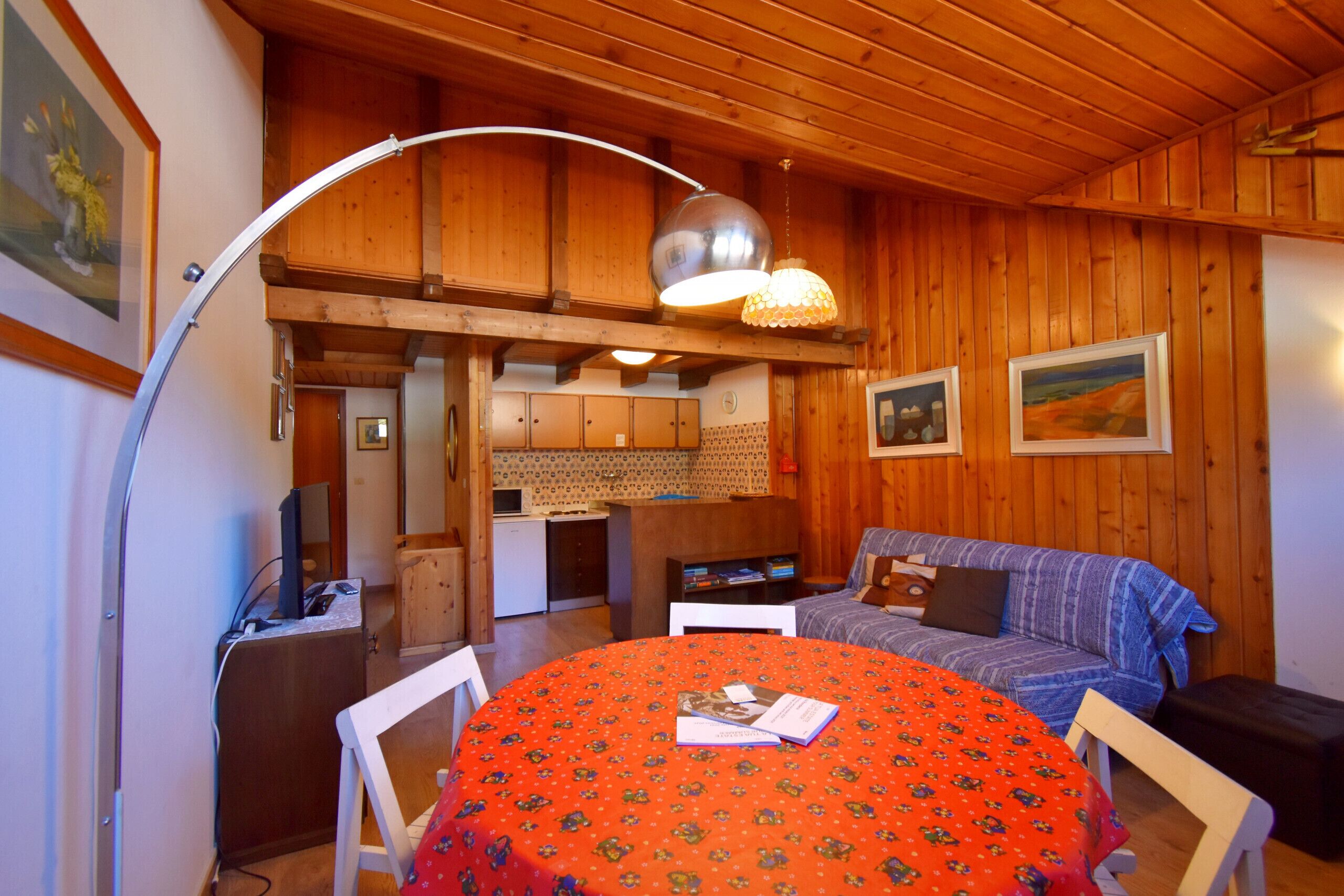 Una habitación acogedora con techo de madera, una mesa de comedor con mantel rojo, un sofá y una zona de cocina al fondo.