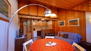 Een gezellige kamer met een houten plafond, een eettafel met een rood tafelkleed, een bank en een keuken in de achtergrond.