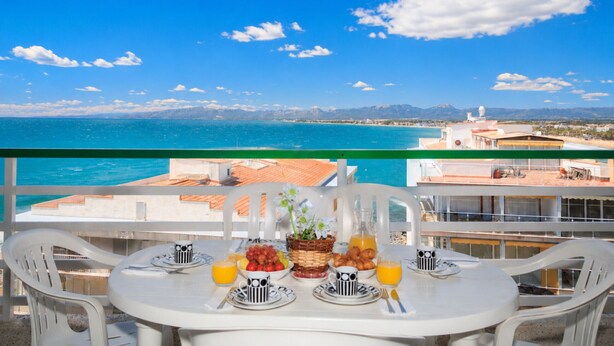 Ein Balkon mit einem Tisch, Frühstücksleckereien, weißen Plastikstühlen und Blick auf das Meer und die Berge.