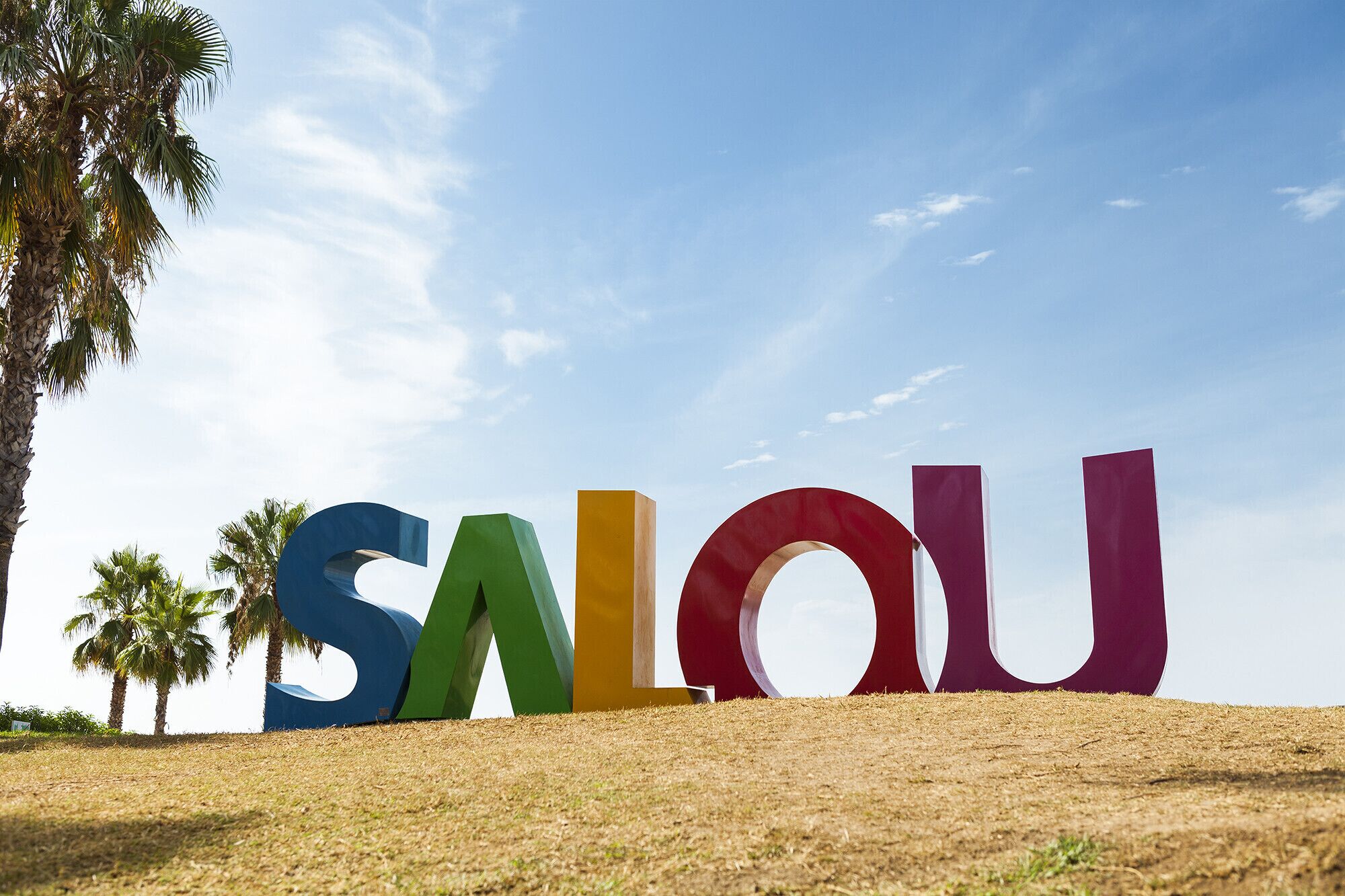 Large, colorful letters spelling "SALOU" on a grassy field with palm trees in the background.