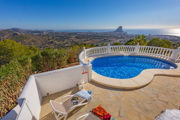 Ein privater Pool mit weißer Brüstung, einem Tisch und einem Stuhl mit Blick auf eine Küstenstadt.