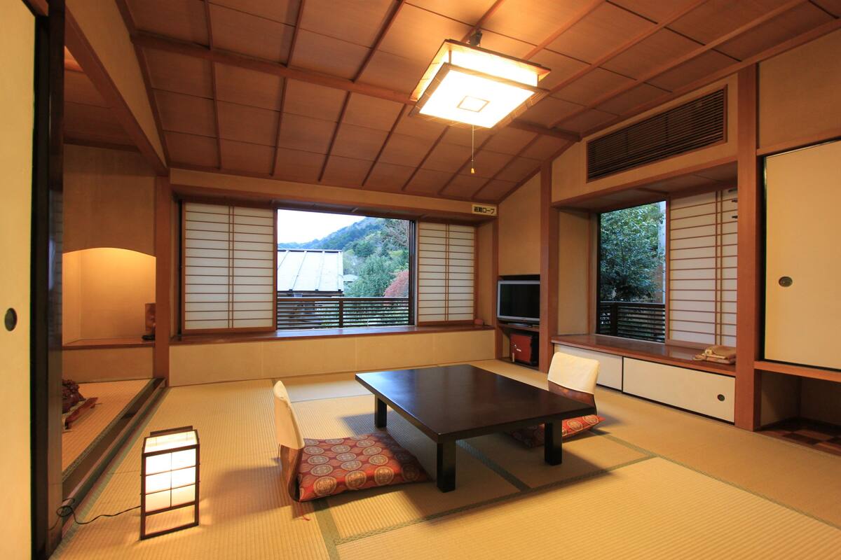 A traditional Japanese room with tatami flooring, a low table, cushions, sliding shoji doors, and a television.
