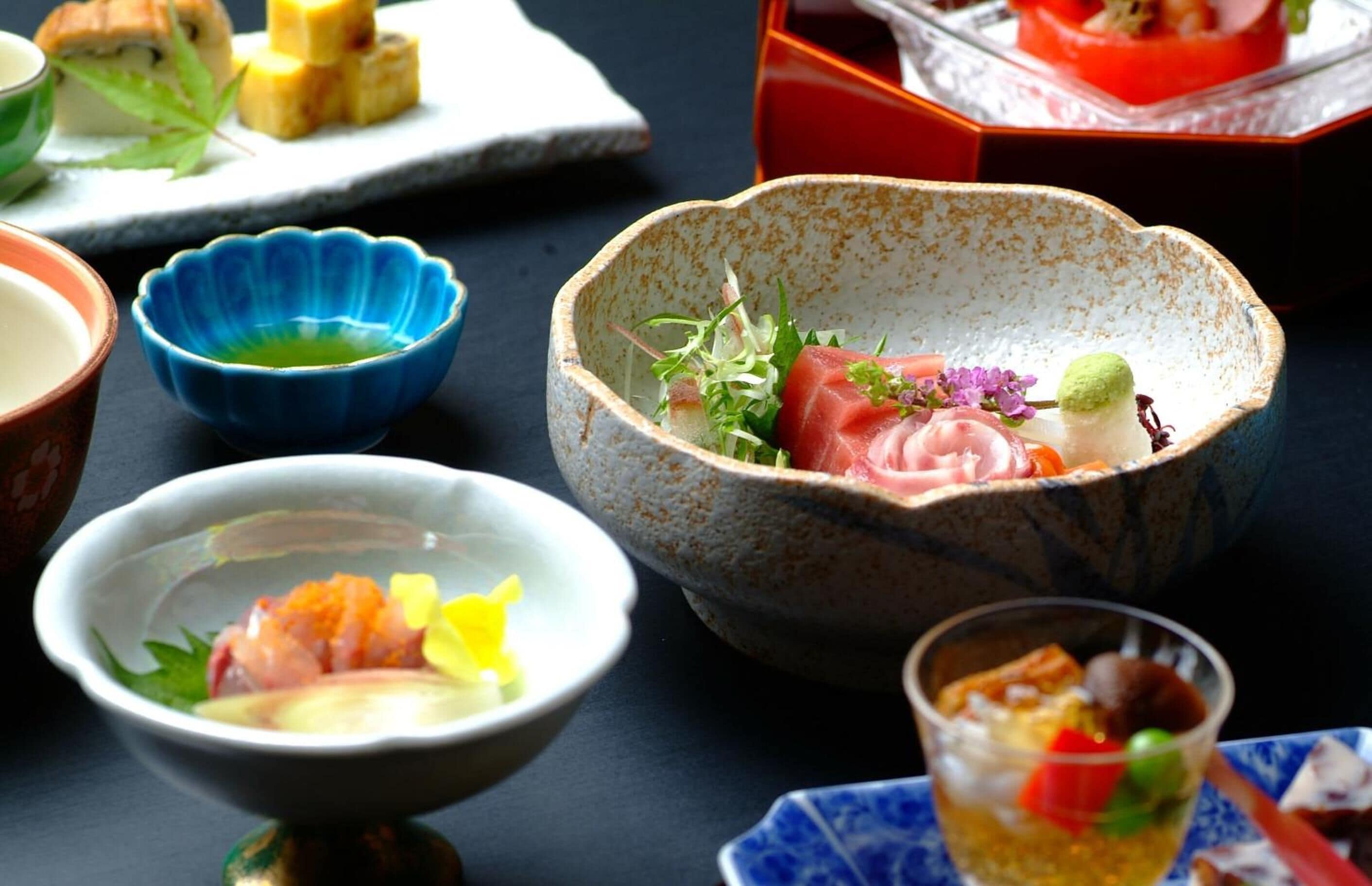 Diversos platos en cuencos tradicionales, como sushi y sashimi, sobre una mesa oscura.