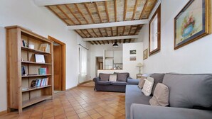 Un salon avec un canapé, une bibliothèque, un sol carrelé, une cuisine à l’arrière-plan et un plafond avec des poutres apparentes.