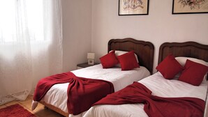 A hotel room with two beds, each with white bedding and red pillows. There is a wooden headboard, a bedside table with a lamp, and a red rug on the floor.