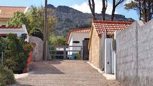 A gravel driveway leading to a gated entrance of a building with a tiled roof, surrounded by greenery and mountains in the background.