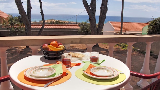 A table set with plates, bowls, a fruit bowl, glasses, and cutlery on a balcony overlooking the sea.