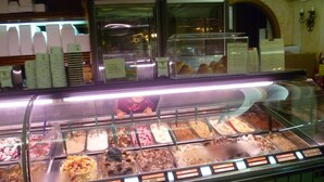 Vitrine com sobremesas, diversos sabores de sorvete e uma pessoa atrás do balcão.