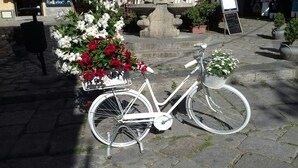 Uma bicicleta branca com cesto e um cesto da frente cheio de flores, estacionada em calçada de pedra.
