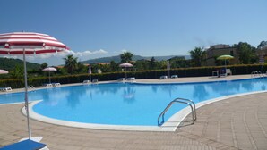 Piscina com espreguiçadeiras e guarda-sóis, cercada por área ajardinada com sebes e palmeiras.