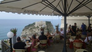 Restaurante ao ar livre com mesas e cadeiras, com vista para a costa e um edifício histórico em um penhasco.