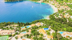 Une vue aérienne d’une zone côtière, avec une mer bleue et limpide, une petite île et une zone résidentielle densément construite, comprenant des maisons et des piscines.