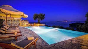 Un espace aménagé au bord de la piscine, avec des chaises longues et des parasols, un mur en pierre et une vue sur la mer au crépuscule.