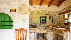Une cuisine rustique avec un mur en pierre, une chaise en bois, un four en briques et une fenêtre donnant sur un paysage verdoyant.