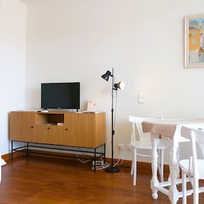Una habitación con un mueble de televisión de madera, una lámpara de pie, una mesa blanca y sillas.