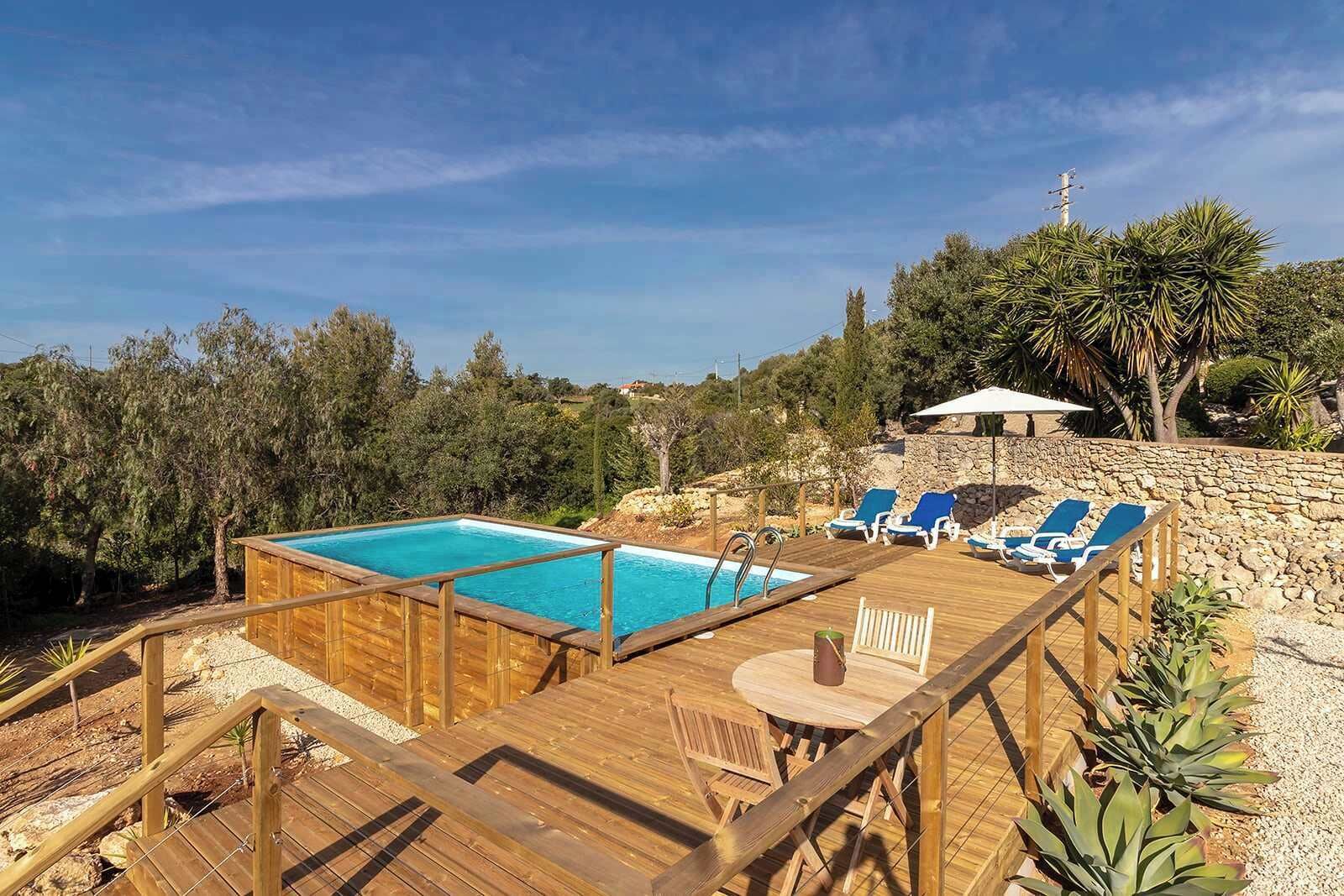 A wooden deck with a pool and lounge chairs, surrounded by trees and a stone wall.