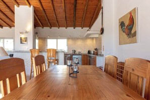 A spacious dining area with a wooden table and chairs, a kitchen in the background, and a rooster painting on the wall.