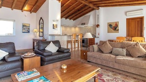 A living room with a wooden ceiling, a fireplace, a kitchen in the background, and a dining area with wooden furniture. - Villa Diane, Luxury Cottage, Ocean views, pool (Benagil)
