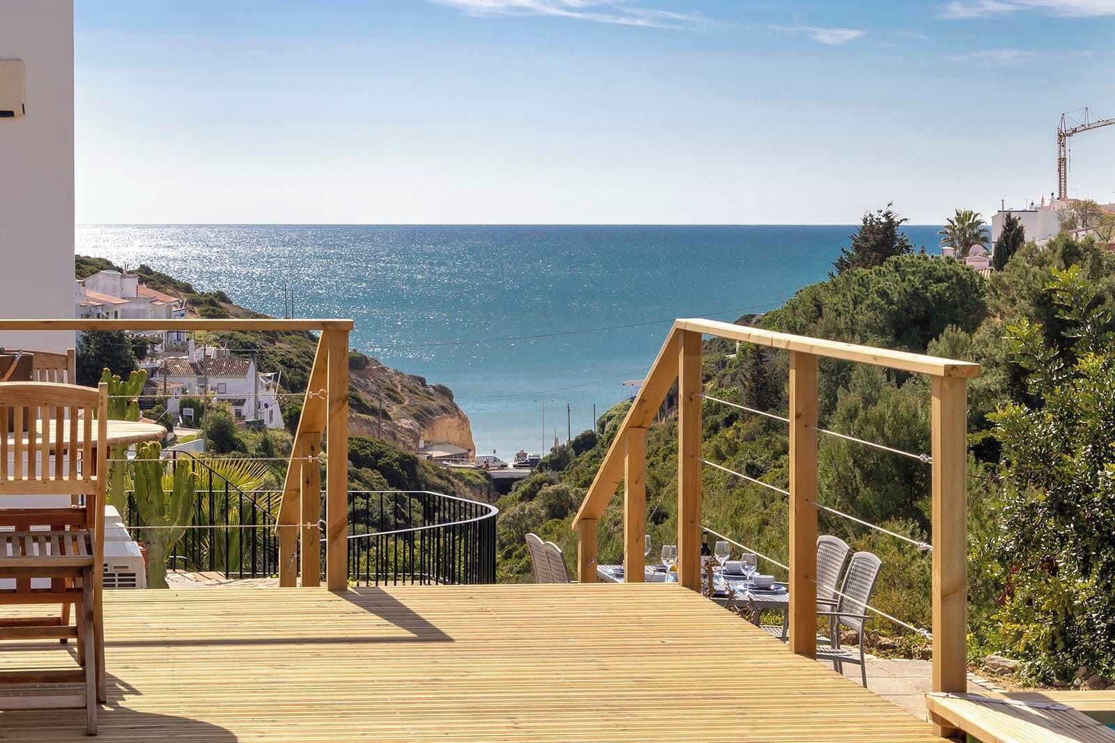 A wooden deck with a railing, overlooking a coastal area with buildings and a clear view of the sea.