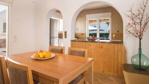 Ein Essbereich mit Holztisch und Stühlen, einer Obstplatte und Blick auf die Küche sowie den Außenbereich durch einen Bogen.