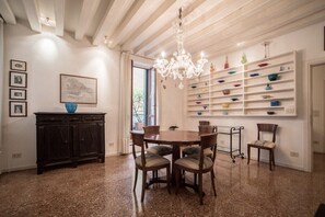 A dining area with a round table, chairs, a chandelier, a cabinet, and a wall-mounted shelf with various items.