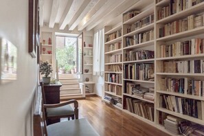 A room with a wooden floor, a bookshelf filled with books, a window with curtains, and a small table with a chair. - Accademia Terrazza (Venice)