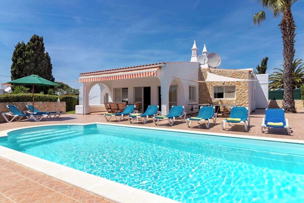 A swimming pool with lounge chairs, a stone building with a tiled roof, and a palm tree.