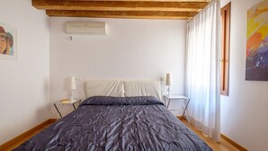 A bedroom with a bed, a nightstand, a lamp, an air conditioning unit, and a window with curtains. - Lion's 1 (Venice)