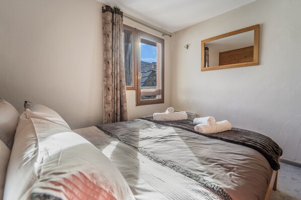 Un llit ben fet amb un estovat estampat, una finestra amb cortines i un mirall amb marc de fusta a la paret.