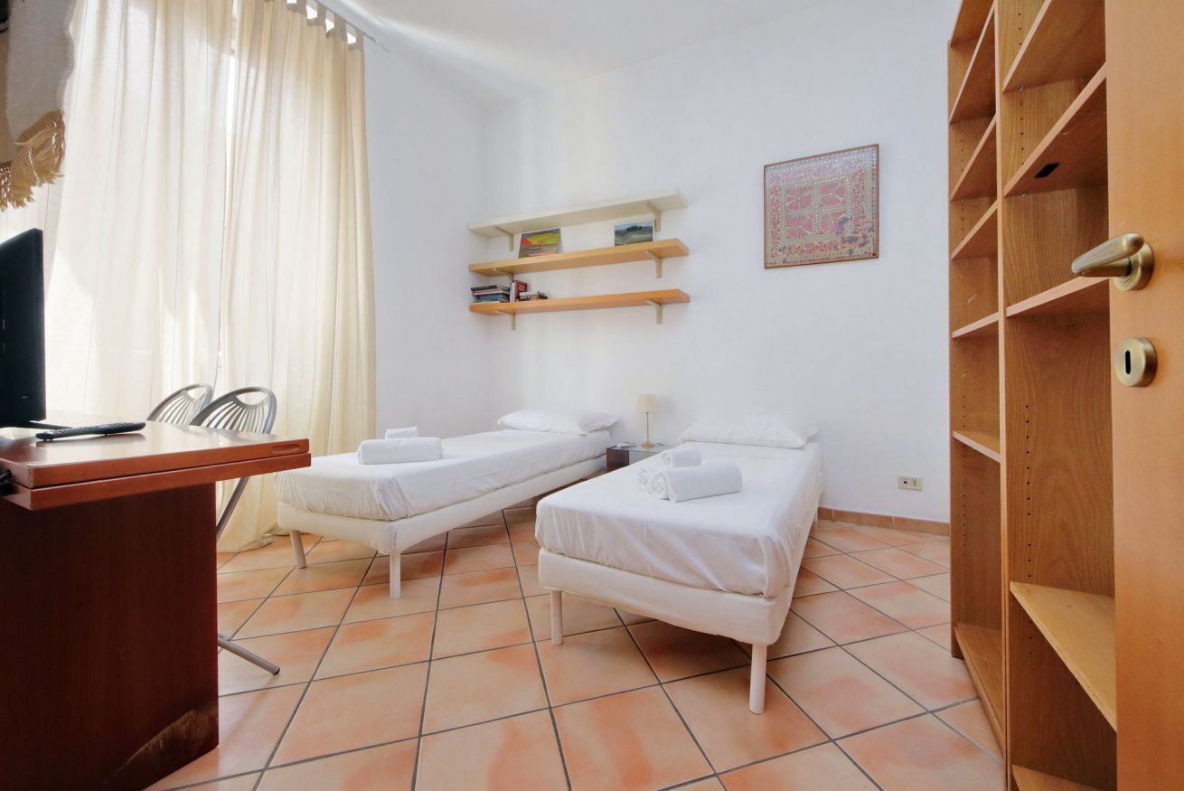 A room with two single beds, a desk with a chair, a wooden wardrobe, and a wall-mounted shelf with books.