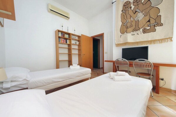 A hotel room with two beds, a desk with a chair, a bookshelf, and a television.